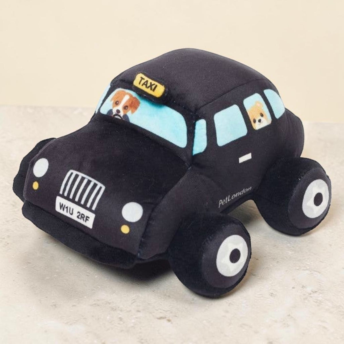 A plush toy shaped like a black taxi car with light blue windows, featuring dog faces in the windshield and side windows, photographed on a light beige surface.