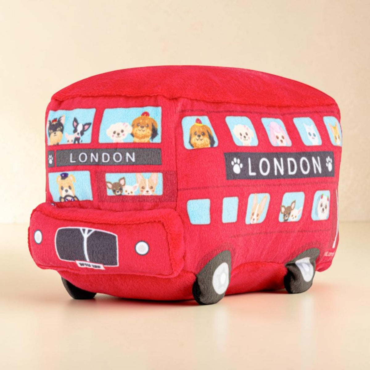 A red plush toy shaped like a double-decker London bus, featuring cartoon animal characters in the windows and 'LONDON' text on the side, photographed against a plain beige background.