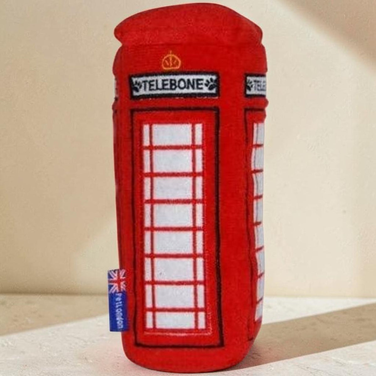 A red plush toy shaped like a classic British telephone booth, featuring white window panes, a black sign reading 'TELEBONE', and a small blue fabric tag near the bottom.