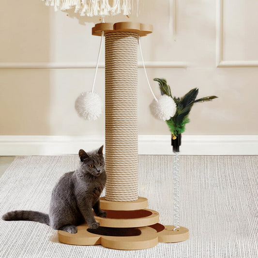 A gray cat sits beside a wooden cat tree with rope-wrapped posts and hanging white pom-poms, while a feather toy dangles nearby in a styled indoor room setting.