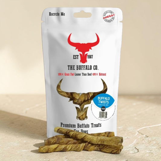 A stand-up pouch of The Buffalo Co. Premium Buffalo Treats, with several twisted dog treats placed in front on a light stone surface.