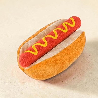 A plush hot dog pillow shaped like a sausage in a bun, with red fabric for the sausage, yellow fabric for the mustard, and tan fabric for the bun, shown on a light beige background.