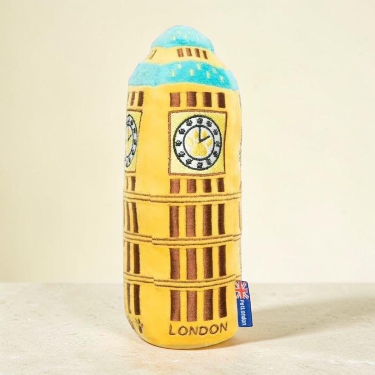 A yellow London Tower of London plush toy with a blue dome top, clock face details, and a small blue tag on the side, set against a plain beige background.