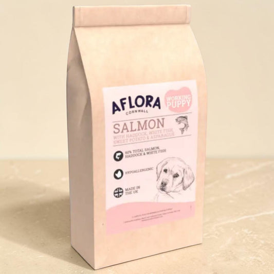 A bag of Aflora Cornwall Working Puppy Salmon dog food, featuring a light pink paper pouch with a white and pink label, set against a neutral beige background.
