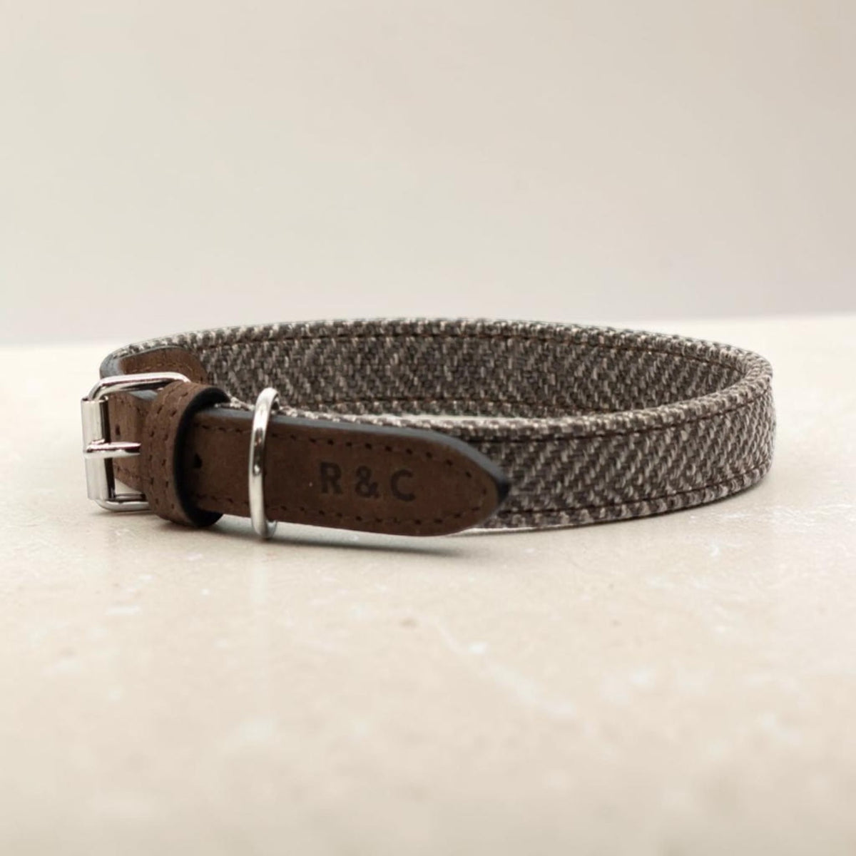 A woven dog collar with a brown leather strap and silver-tone buckle, resting on a light beige surface. The collar features a textured gray and brown herringbone pattern, and the inside leather tab is stamped with 'R&C'.