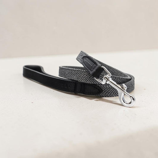 A gray and black herringbone patterned dog leash with a black leather handle and silver metal clasp, resting on a light beige surface.