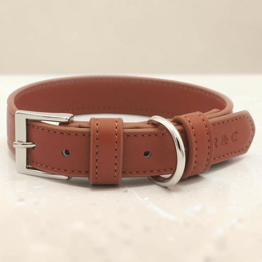 A brown leather dog collar with a silver-tone metal buckle and D-ring, displayed on a light beige textured surface.